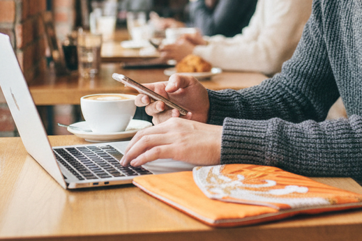 カフェのテーブルでノートパソコンとスマートフォンを操作する手元とオレンジ色のPCスリーブ